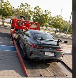 سطحة هيدروليك حديثة من أسطول سطحة جدة تحمل سيارة BMW M8 رياضية فارهة ذات جناح خلفي كبير، متوقفة بأمان في شارع مشجر في حي المحمدية بجدة. الخدمة متاحة على مدار الساعة لنقل السيارات الرياضية والفاخرة في المنطقة